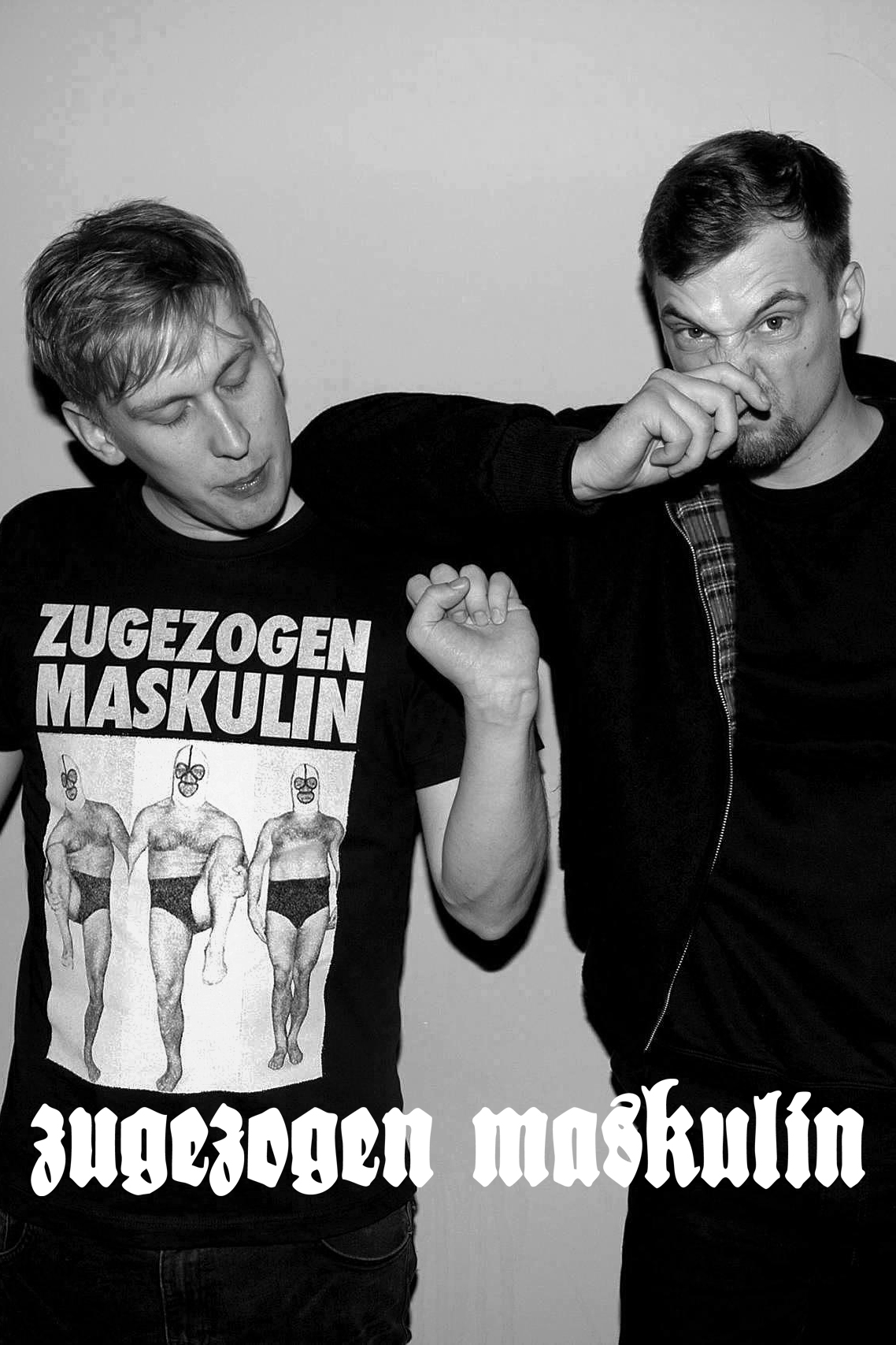 Zwei junge Männer, einer trägt ein T-Shirt mit der Aufschrift 'Zugezogen Maskulin' und drei Wrestlerfiguren, der andere hält sich die Nase zu
